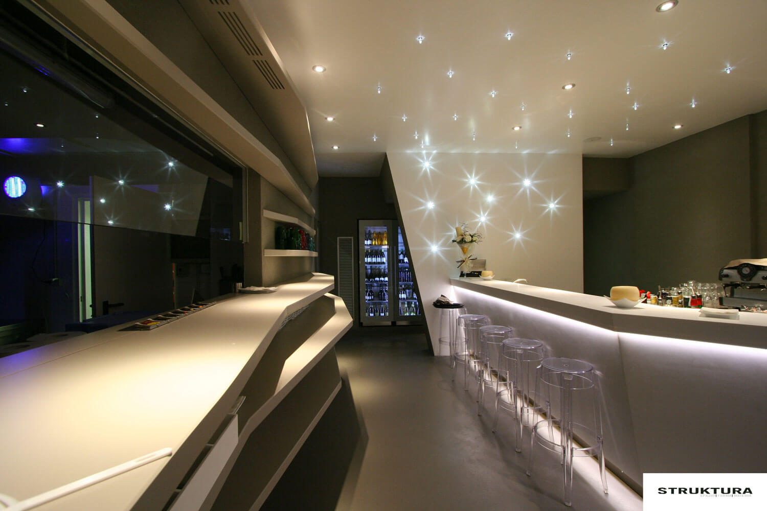 Moderne Bar mit fugenloser Oberfläche, stimmungsvollem Licht und transparenten Barhockern.